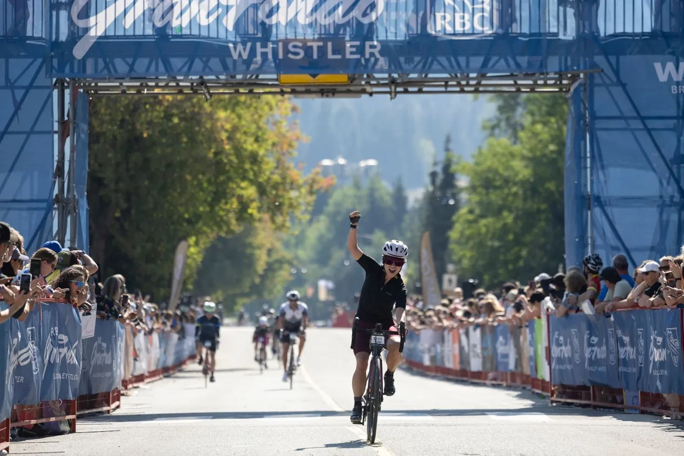 A cyclist raises an arm in victory while crossing the finish line at the RBC GranFondo Whistler, with spectators cheering along both sides of the road.