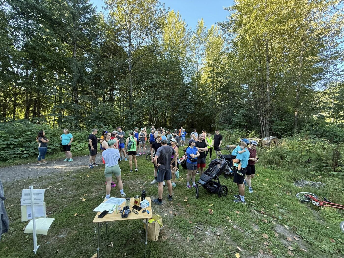 A group of runners, walkers, and families with strollers gather at the start of the Mamquam Spawning Trail parkrun in Squamish, surrounded by tall trees and greenery on a sunny morning.